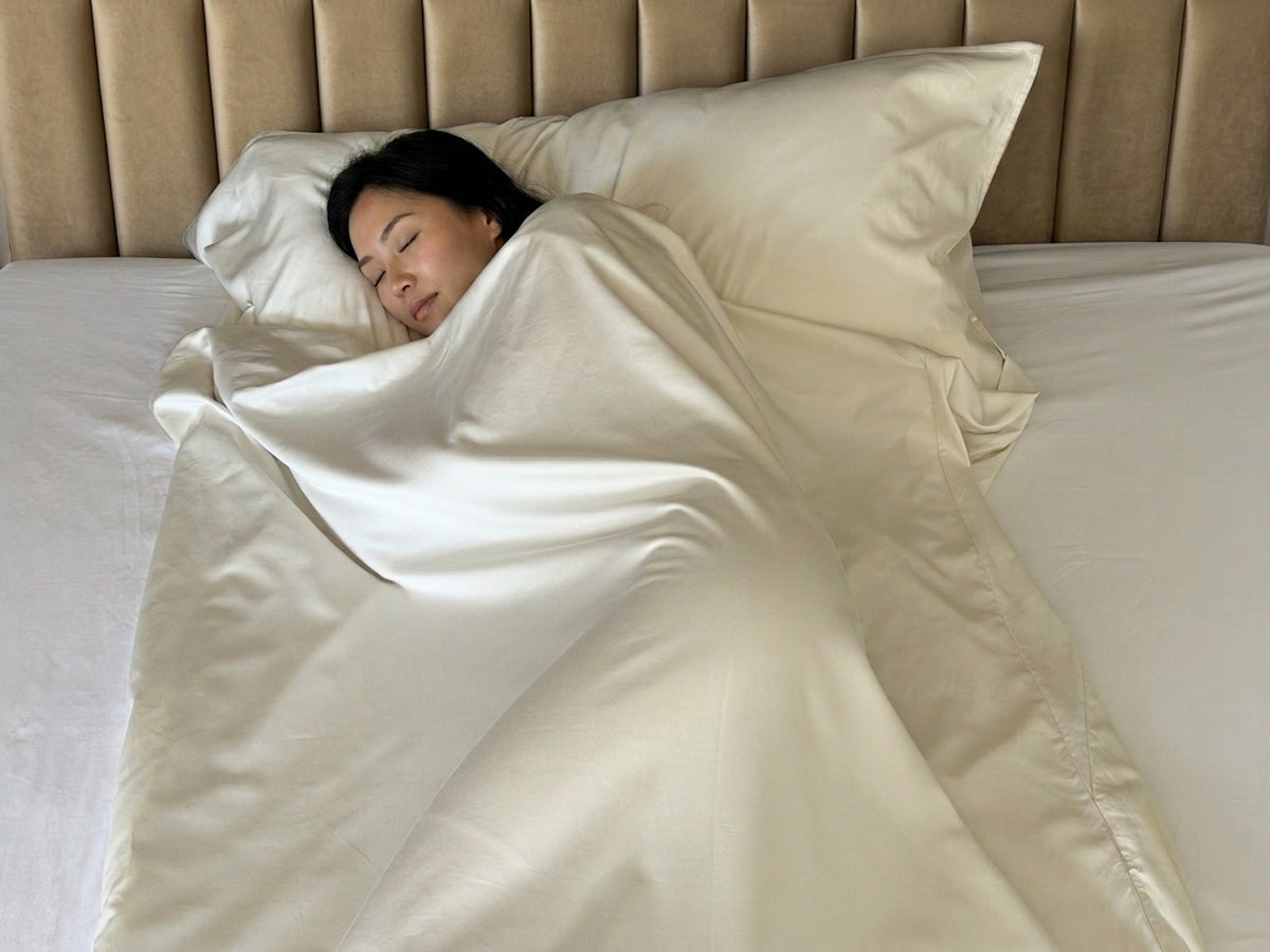 Woman sleeping peacefully wrapped in the UpsideLuxe Sleep Sack, a cream Supima cotton sateen travel bedding layer, against an upholstered tan headboard
